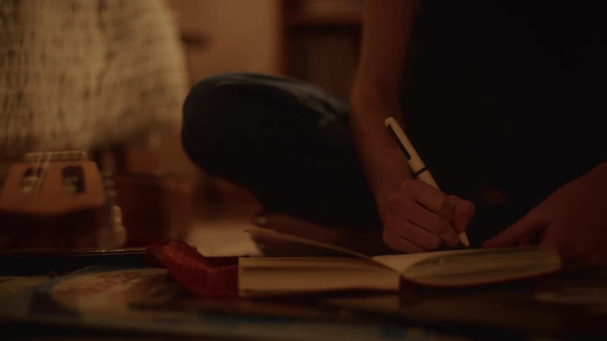 Close-up shot of a person sitting on the floor writing in a journal in a cozy, dimly lit room at night