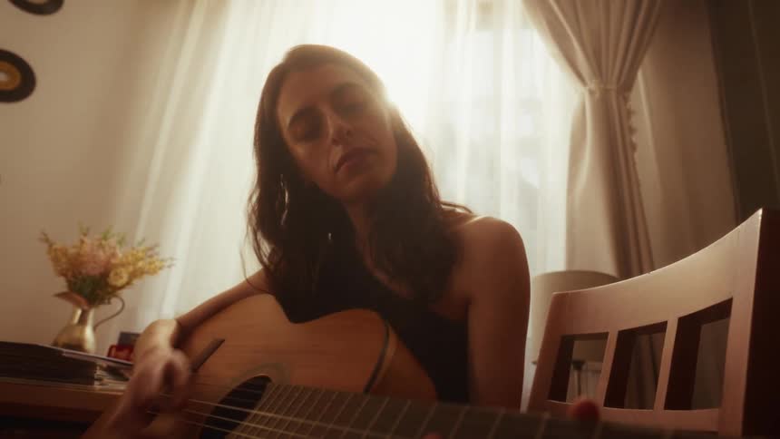 A young woman emotionally plays her acoustic guitar, silhouetted by the warm sunlight from a window
