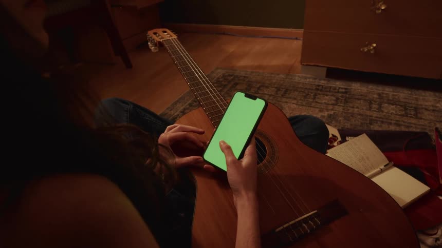A person with an acoustic guitar on their lap scrolls through a smartphone with a green screen in a cozy room