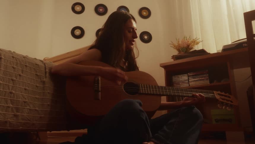 Happy young woman sings and plays an acoustic guitar while sitting on the floor of her cozy bedroom at night