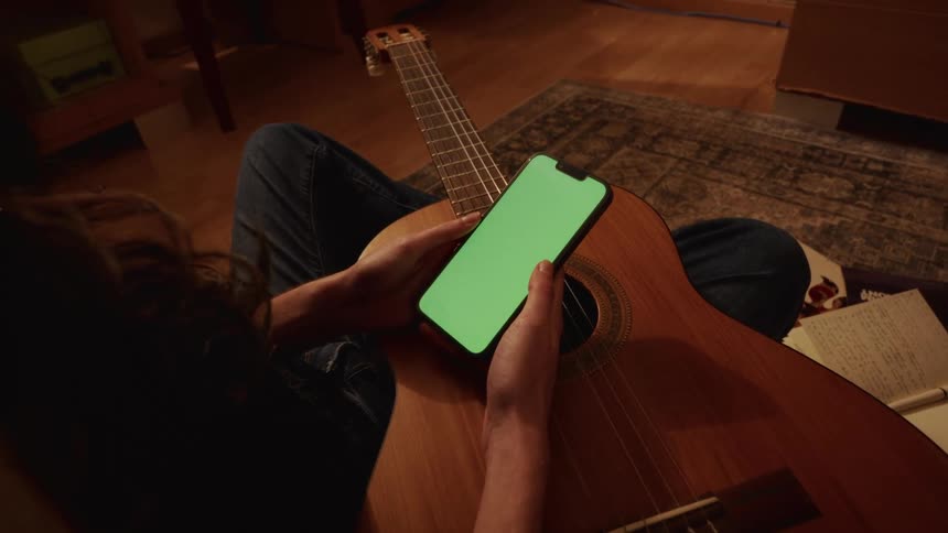 Over-the-shoulder shot of a musician holding an acoustic guitar while scrolling on a smartphone with a green screen