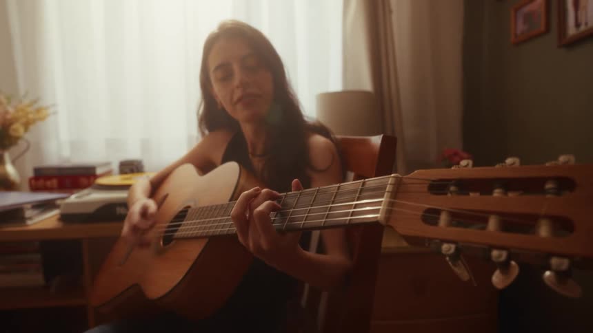 Close up of a smiling woman playing an acoustic guitar in her cozy room with warm natural light