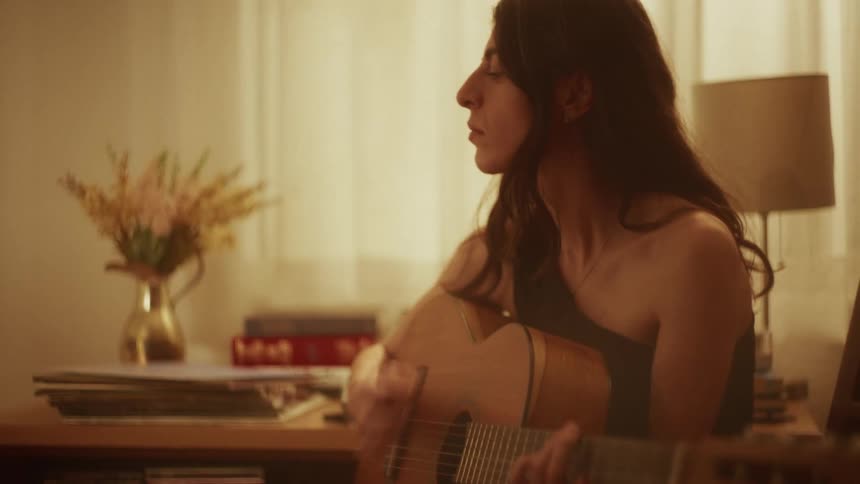 An introspective young woman plays a soft melody on her acoustic guitar in a warm, dimly lit room