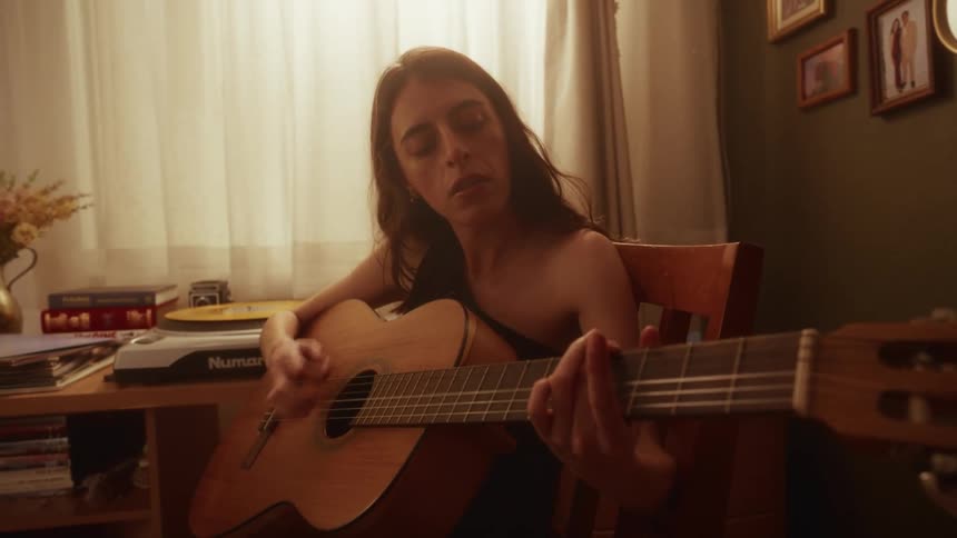 A focused young woman sits alone in her warmly lit room, strumming and playing a song on her acoustic guitar