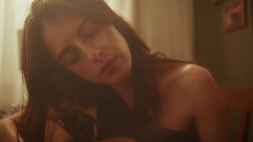 A close-up shot of a soulful woman with her eyes closed as she plays the acoustic guitar in a warmly lit room