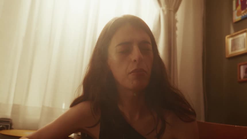 An intimate close-up shot of a woman with her eyes closed, passionately singing and playing acoustic guitar in a warm, hazy room