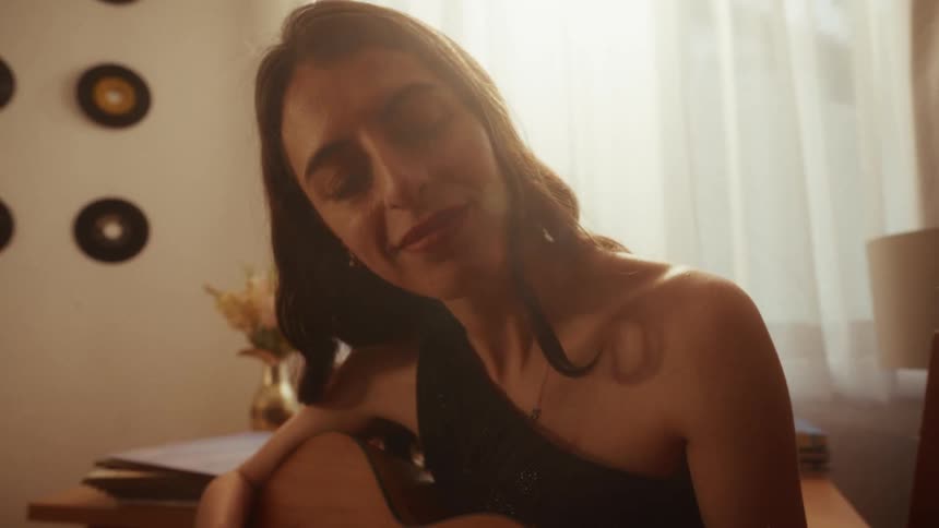 A woman plays an acoustic guitar with emotion and her eyes closed, bathed in the warm light from a nearby window
