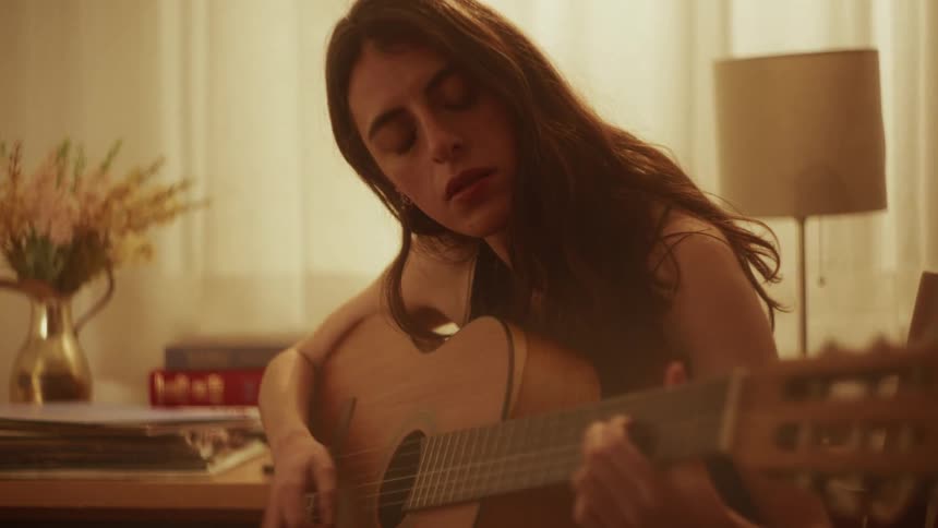 A pensive young woman with her eyes closed emotionally plays a song on an acoustic guitar in a warmly lit room