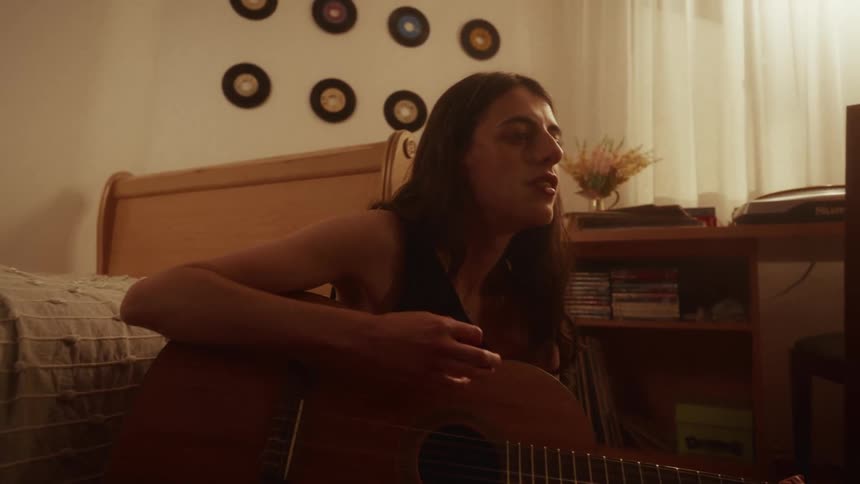 A young woman passionately plays the acoustic guitar and sings in her cozy bedroom at night, surrounded by her vinyl record collection