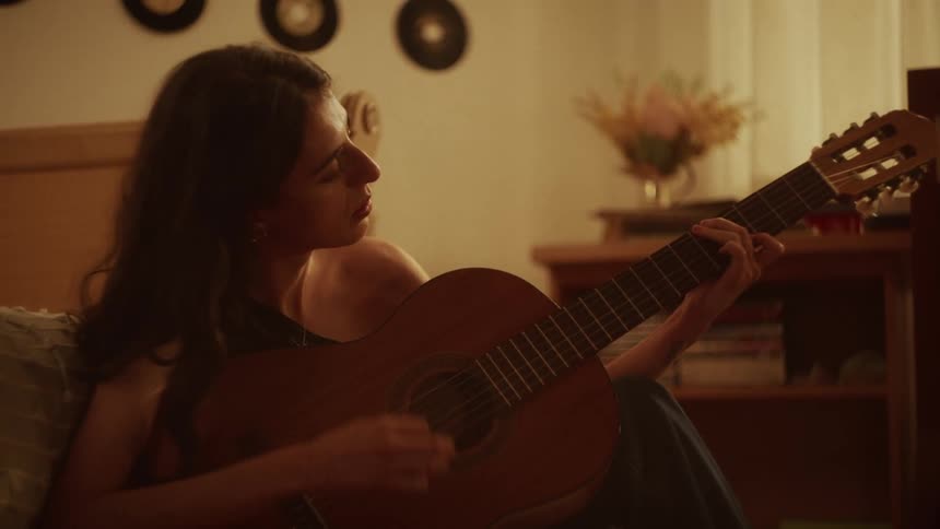 A young woman sits on her bed in a warmly lit room and plays an acoustic guitar