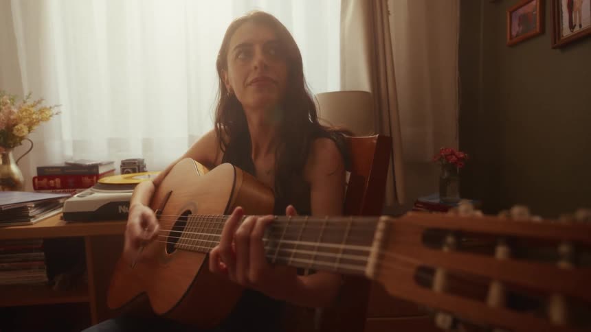 A talented young woman serenely plays her acoustic guitar in a warm, sunlit room at home