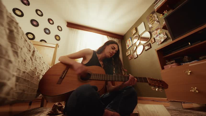 A low angle wide shot of a young woman passionately playing an acoustic guitar while sitting on the floor of her cozy, retro-decorated bedroom