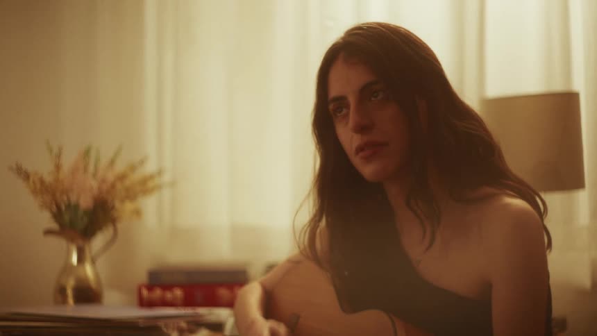 A soulful young woman with long dark hair plays a heartfelt song on her acoustic guitar in a cozy, warmly lit room