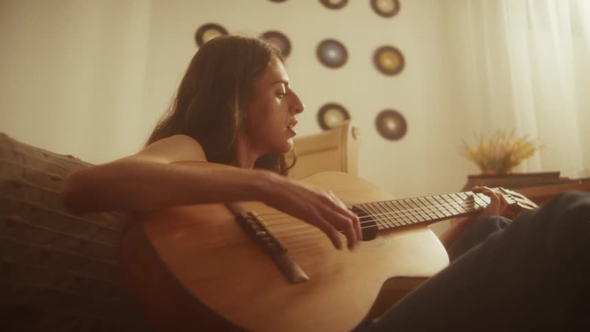 Close up of a young woman singing and playing the guitar in her warmly lit bedroom with vinyl records on the wall