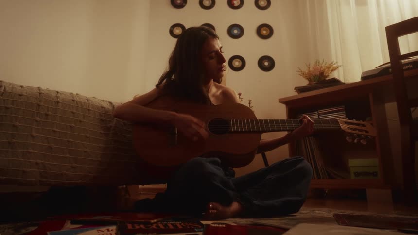A young woman in a dimly lit, cozy room picks up an acoustic guitar in slow motion and begins to play and sing
