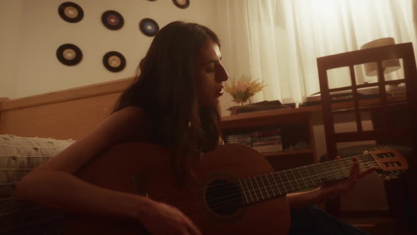 A young woman passionately sings and plays an acoustic guitar in her dimly lit bedroom at night