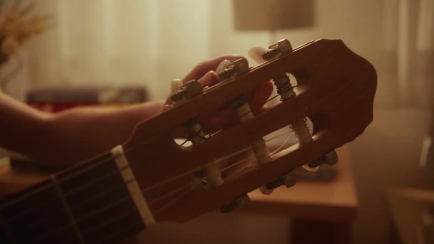 A musician's hand is seen in close-up, delicately tuning a classical guitar in a warm, cozy setting