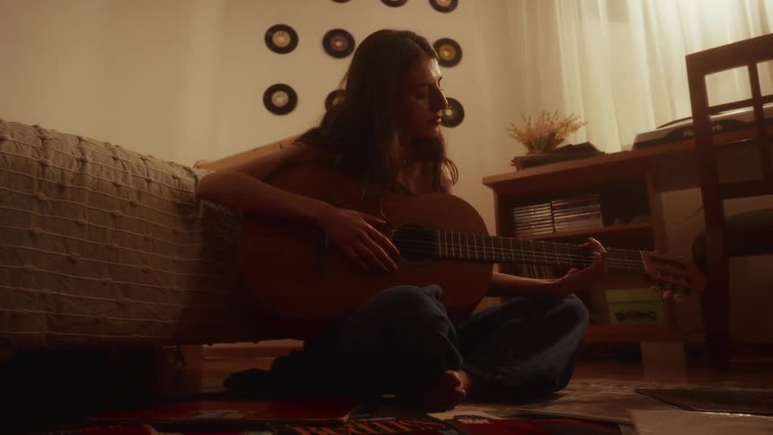 A young woman sits on her bedroom floor surrounded by vinyl records, picks up her acoustic guitar, and begins to play in the warm, ambient light