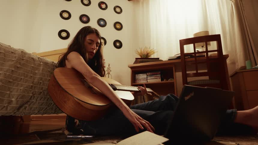 A focused young person takes an online guitar lesson on their laptop while sitting on the floor of their warmly lit bedroom