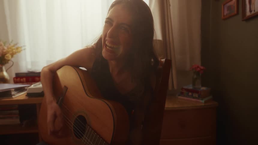 A passionate young woman laughs and sings while playing her acoustic guitar in a warmly lit room