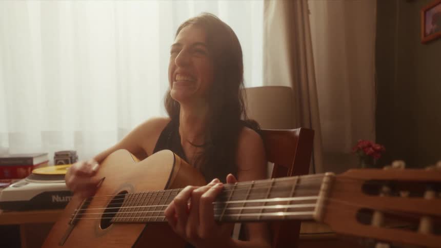 A joyful young woman laughs and sings while passionately playing her acoustic guitar in a sunlit room, captured in slow motion
