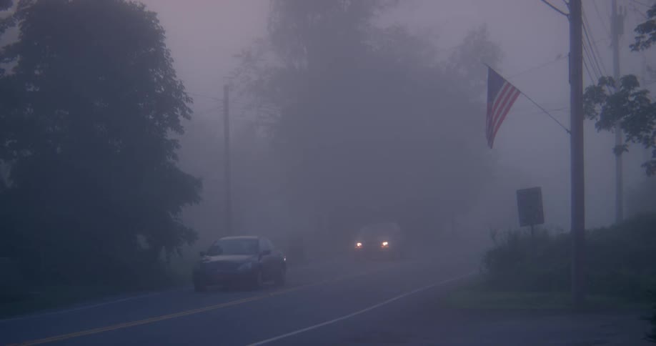 Cars driving down a foggy country road past an american flag