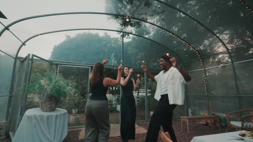 Joyful diverse friends dance energetically with sparklers at an evening party inside a decorated greenhouse