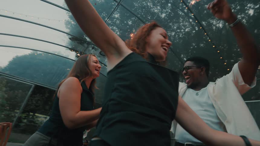 A diverse group of friends laugh and dance together with sparklers at a party in a greenhouse in slow motion