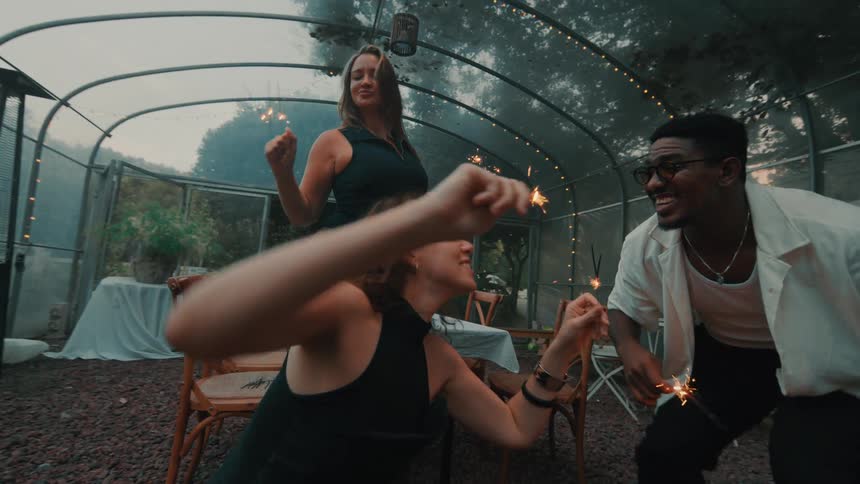 Joyful diverse friends dance in slow motion with burning sparklers at a party inside a stylishly decorated greenhouse