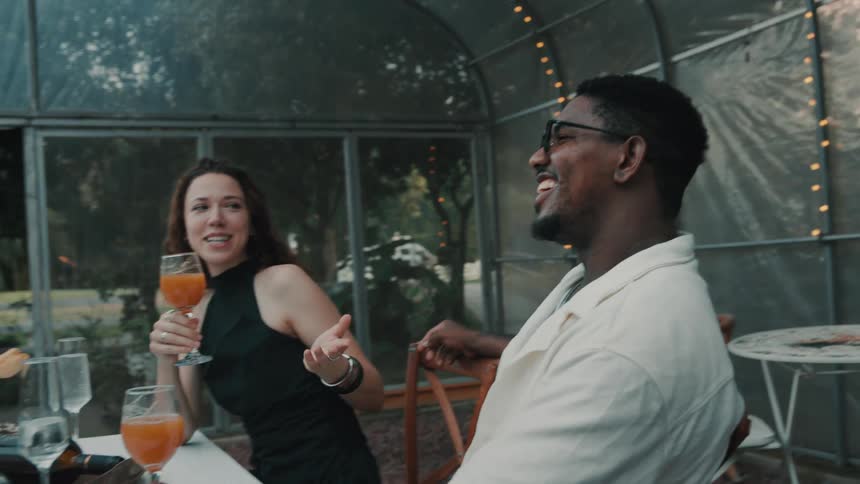 A happy diverse couple laughs and enjoys drinks while sitting at a dinner table in a beautiful greenhouse setting