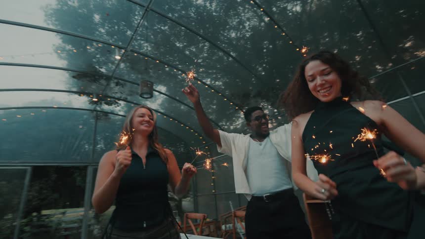 Diverse friends joyfully dance in slow motion with sparklers at an evening party inside a decorated greenhouse