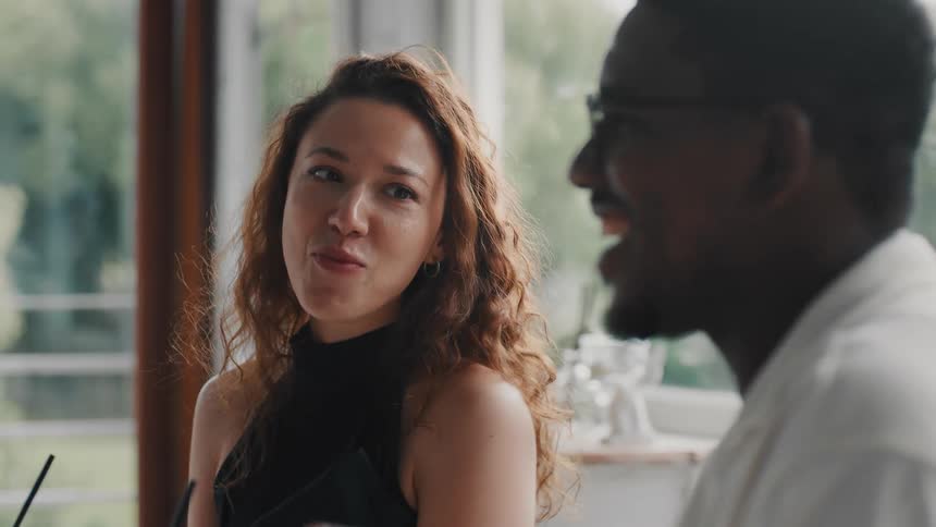 A diverse group of friends smile and engage in lively conversation while sitting at a table in a restaurant during a daytime celebration