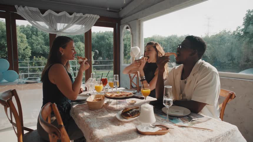 A diverse group of cheerful friends enjoy sharing a pizza and drinks together at a sunlit restaurant