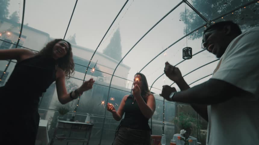A diverse group of happy friends dances with sparklers in slow motion during a celebration in a beautifully lit greenhouse