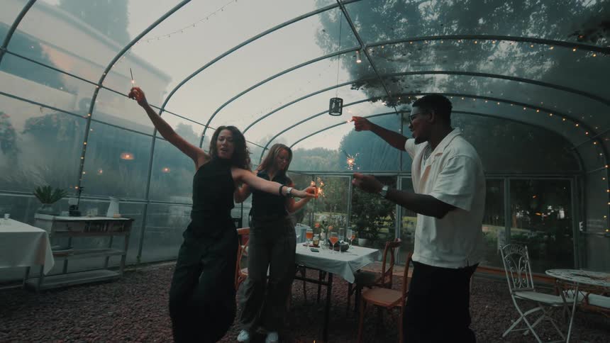 Three happy friends dance with sparklers in slow motion during an evening party in a greenhouse