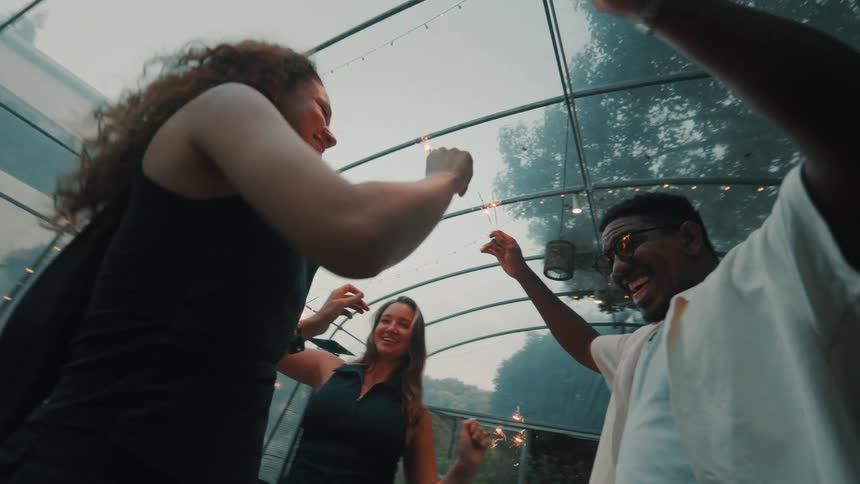 A low-angle slow-motion shot captures three happy, diverse friends laughing and dancing with sparklers at an evening party in a greenhouse
