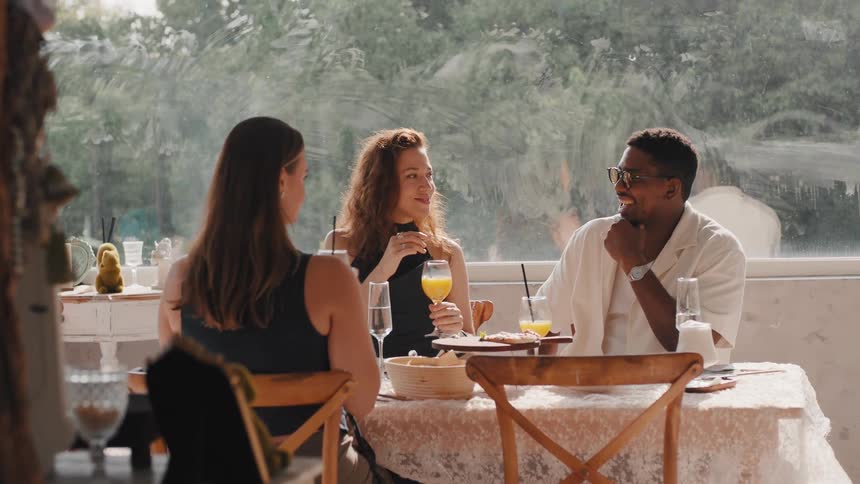 A group of diverse friends enjoy a sunny brunch, laughing and toasting with orange juice on a restaurant patio