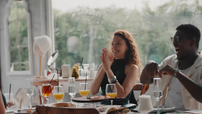 A slow motion shot of happy, diverse friends celebrating with high-fives while dining together in a bright restaurant