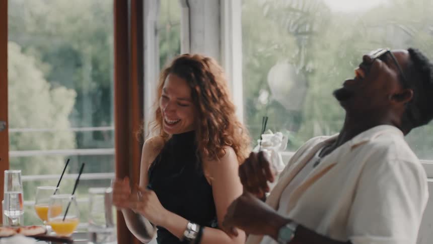 A joyful interracial couple laugh and dance in their seats while celebrating at a restaurant table in slow motion