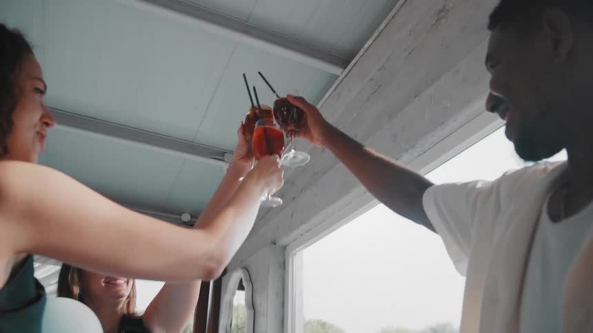 A diverse group of friends happily toast with their orange cocktails in slow motion on a bright day