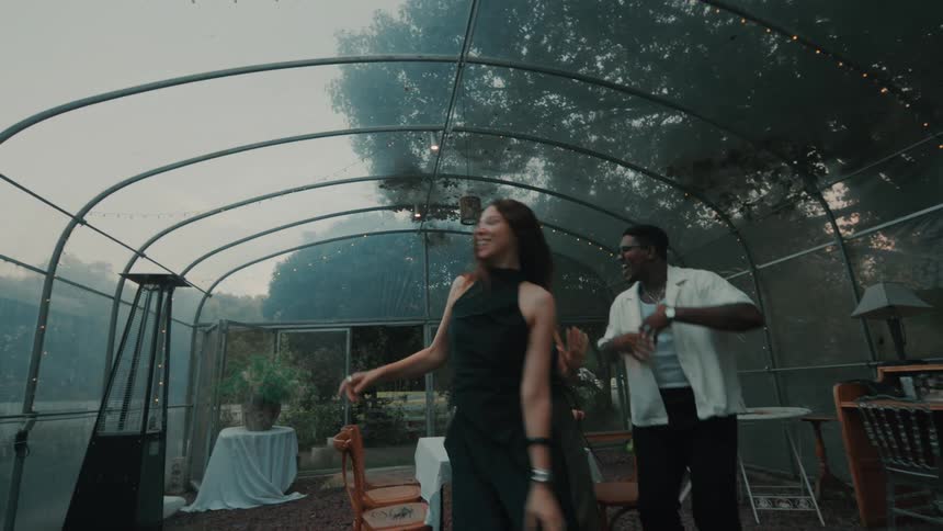 A diverse group of three friends joyfully dance in slow motion inside a stylishly decorated greenhouse during an evening party
