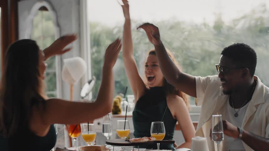 A diverse group of friends laugh and high-five in slow motion while celebrating together at a beautiful restaurant