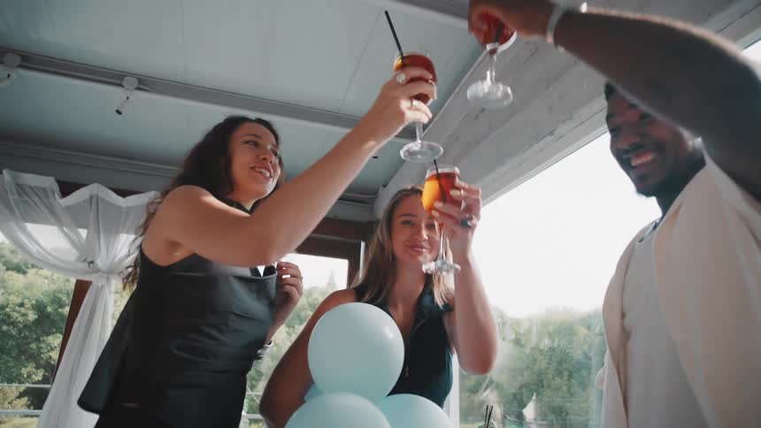 A diverse group of happy friends in slow motion clink their cocktail glasses together to celebrate at a daytime party