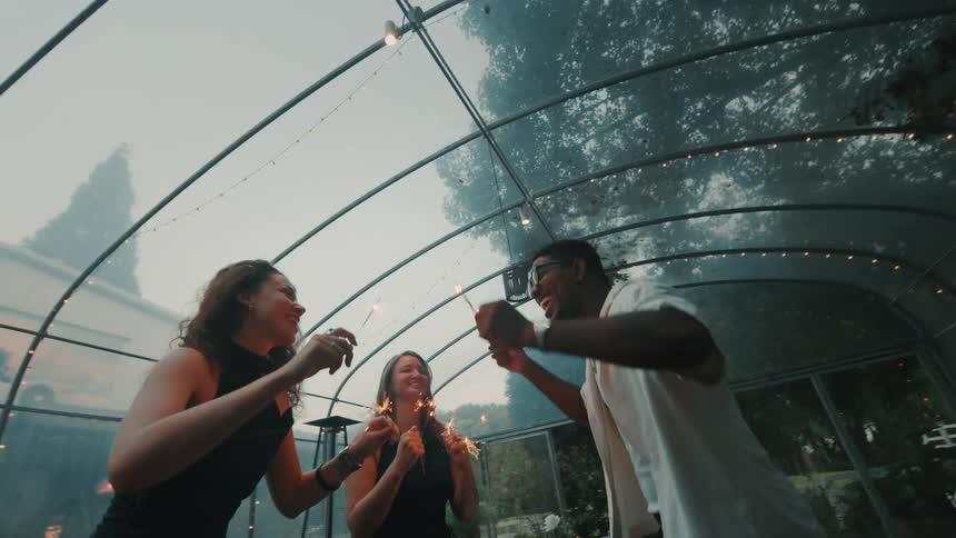 A diverse group of happy friends laugh and dance together with sparklers at an evening party in a modern greenhouse