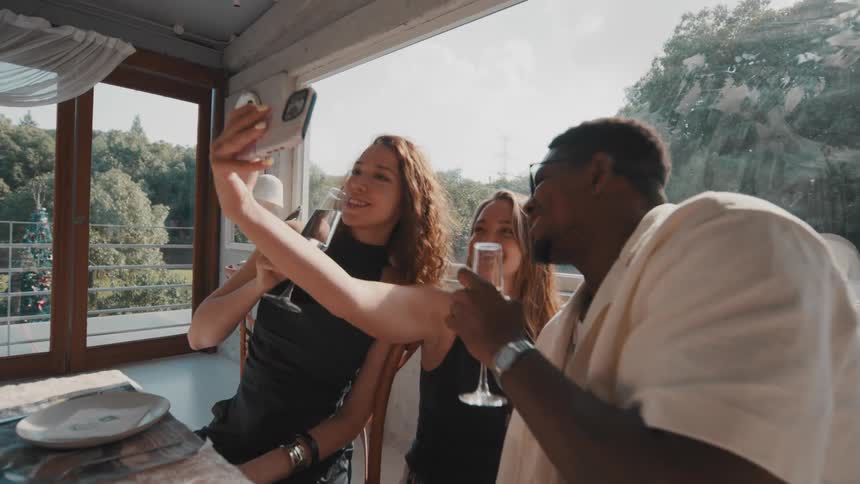 A joyful group of diverse friends capture a happy moment with a selfie, laughing and drinking wine together at a sunny restaurant in slow motion