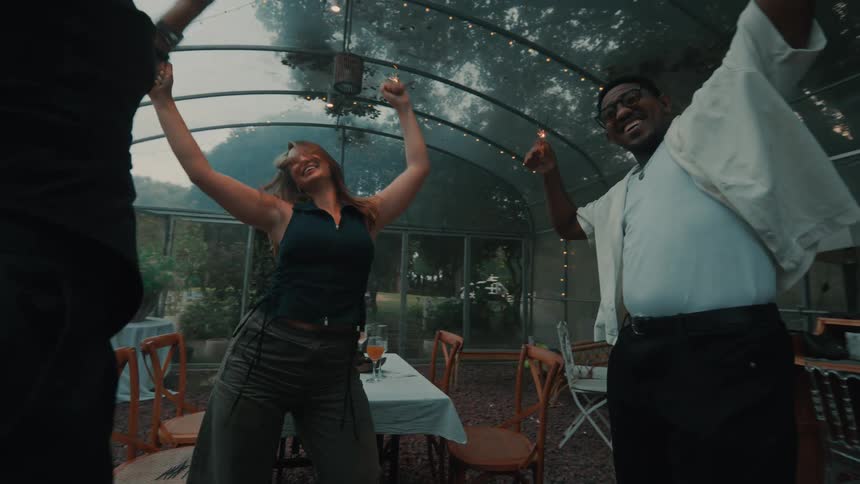 Joyful friends laugh and dance with sparklers at a festive evening party inside a greenhouse in slow motion