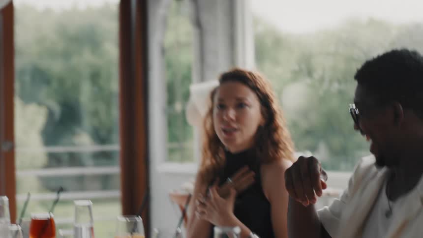 A group of diverse friends joyfully laugh and high-five in slow motion while enjoying a meal together at a sunlit restaurant