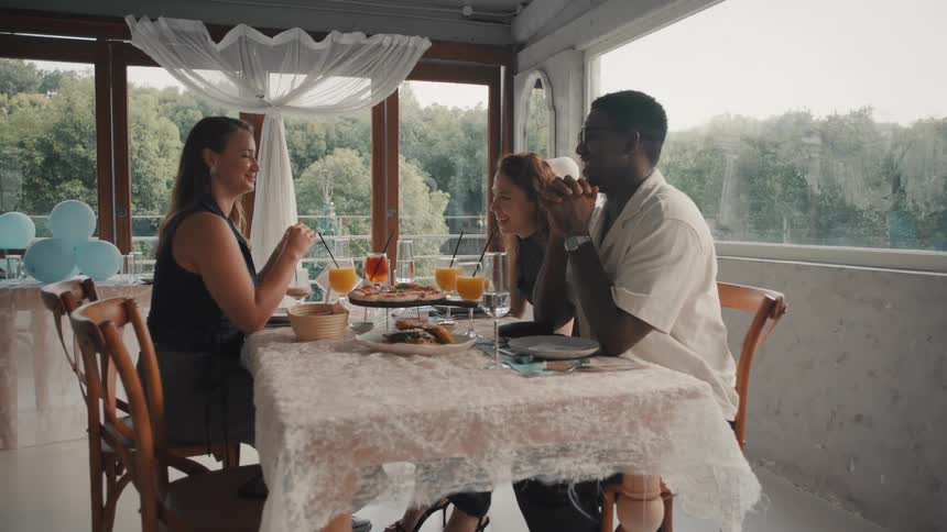 A diverse group of three friends happily celebrates with lunch, laughing and talking at a restaurant table with a scenic view in slow motion