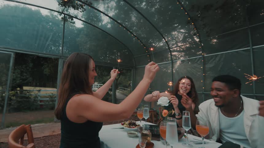 A diverse group of happy friends laugh and celebrate with sparklers at a festive dinner party in a stylish greenhouse