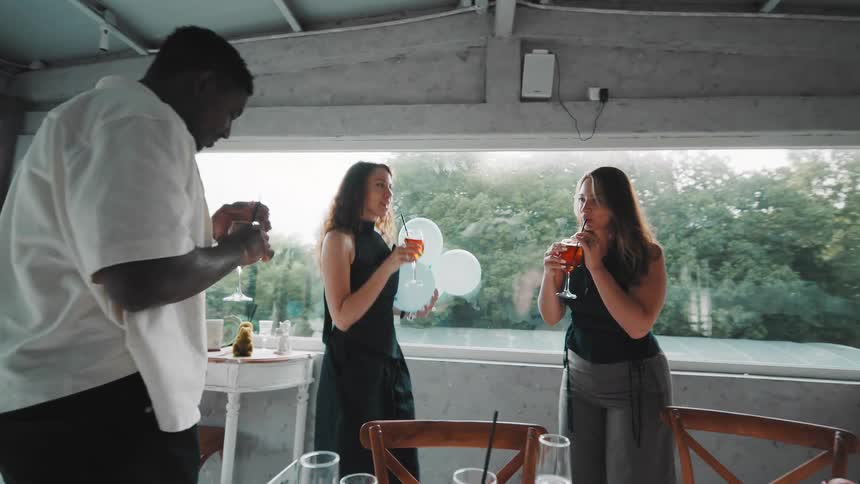 A diverse group of friends in slow motion joyfully toast with cocktails at a celebration in a sunlit restaurant